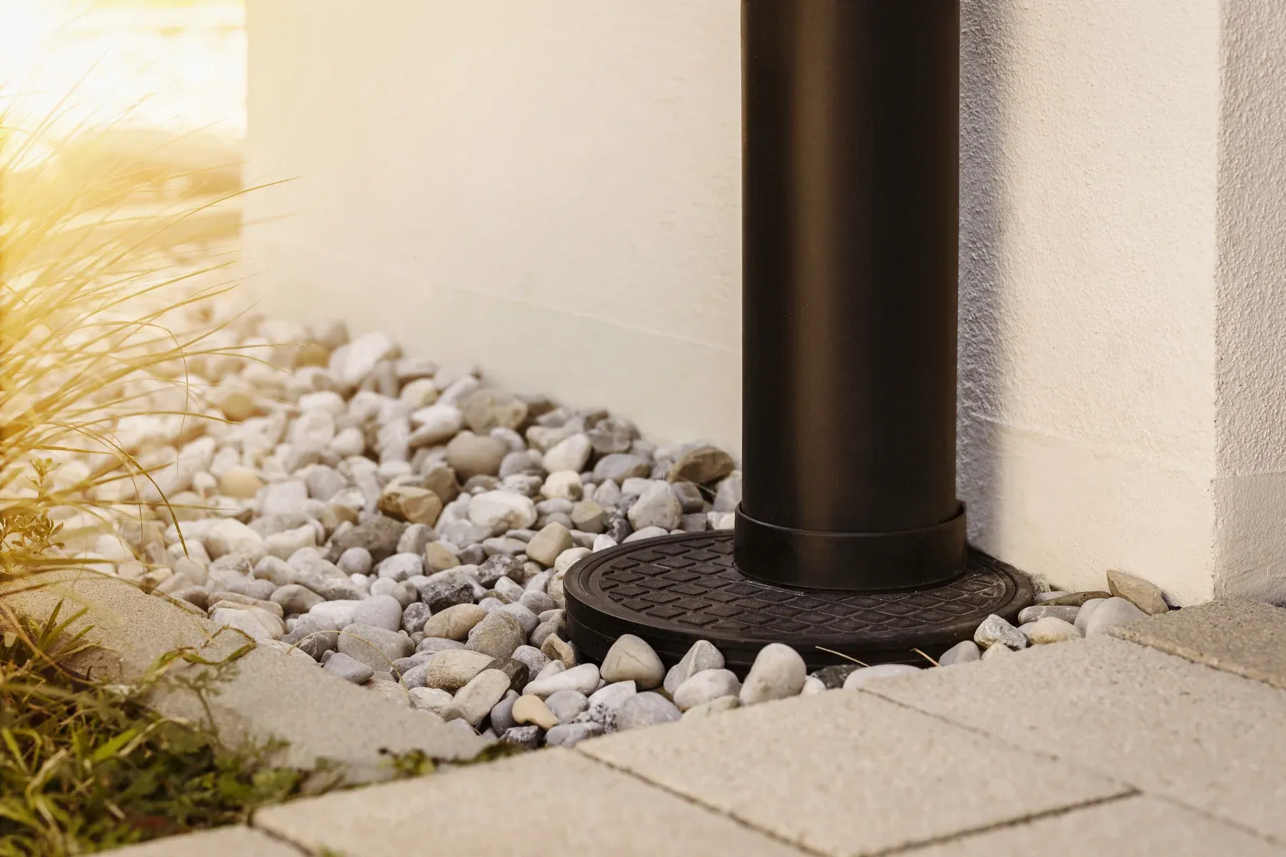 Dark downspout feeding an underground drain inlet surrounded by river rock beside a house wall