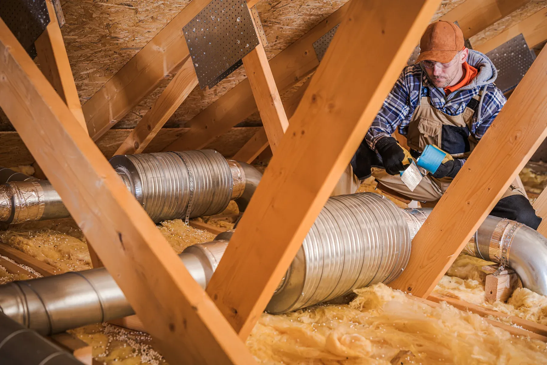 Technician kneeling in an attic with a roll of tape beside silver ducts running between wooden rafters