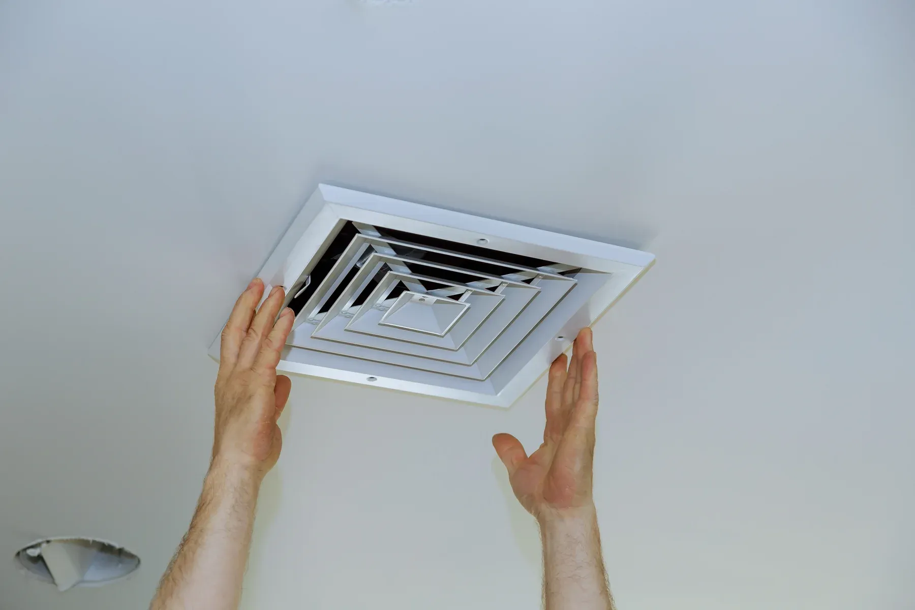 Two hands fitting a white square supply air register into a smooth white ceiling