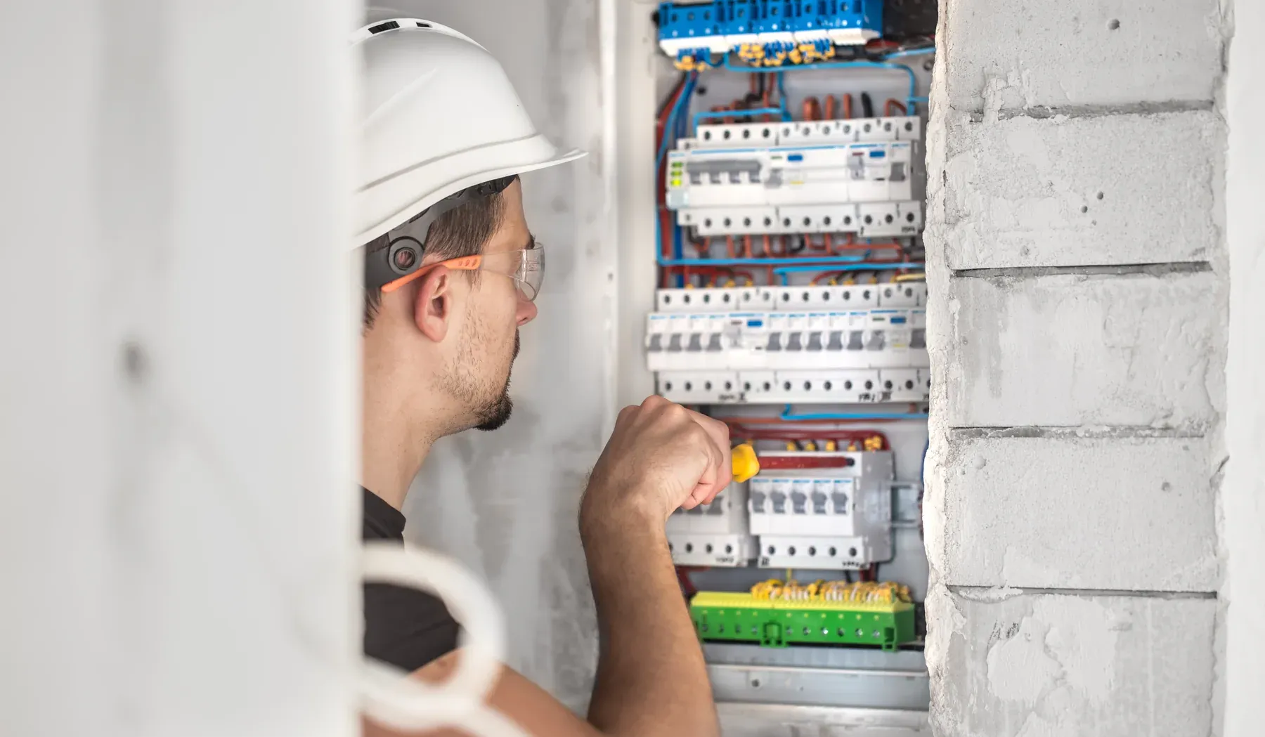 Electrician in a hard hat and safety glasses working inside an open wall-mounted breaker panel