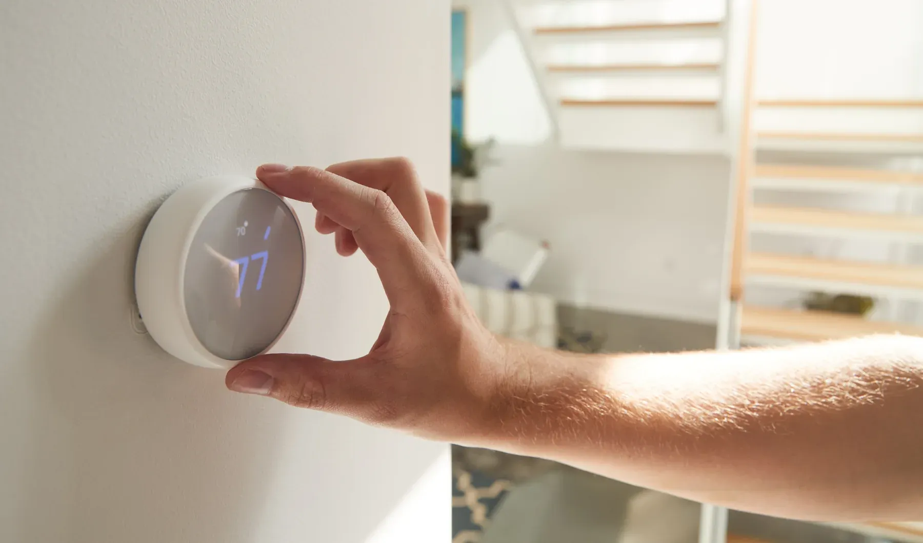 Hand turning the dial of a round white smart thermostat mounted on a light wall